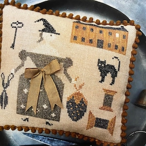 Könnte beinhalten: Ein braunes und beiges besticktes Kissen mit einer Hexe, einem Haus, einer Katze, einer Erdbeere und einer Spule Faden. Das Kissen hat einen braunen Pompon-Besatz.