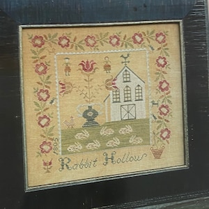 Könnte beinhalten: Eine gerahmte Kreuzstich-Stickerei, die ein Haus mit rotem Dach und einem weißen Lattenzaun darstellt. Die Stickerei ist von einem floralen Rand umgeben und der Text "Rabbit Hollow" ist unter dem Haus gestickt.