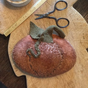 May include: A brown felt pumpkin with a green felt stem and leaves. The pumpkin is sitting on a wooden cutting board with a ruler and a pair of scissors.