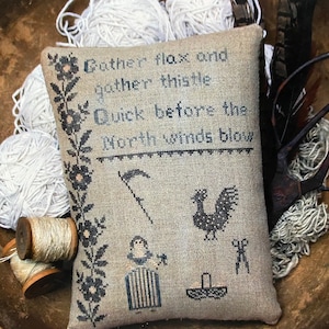 Könnte beinhalten: Ein Kreuzstich-Stickmuster-Kissen mit einem schwarz-weißen Design. Das Kissen zeigt einen Hahn, eine Sense, eine Frau, die einen Korb hält, und einen Korb mit Blumen. Der Text auf dem Kissen lautet "Gather flax and gather thistle Quick before the North winds blow".