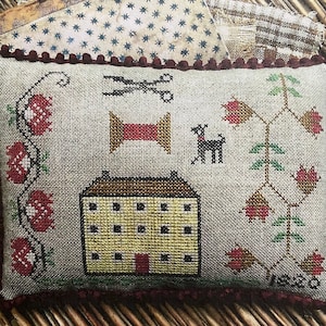 Könnte beinhalten: Ein rechteckiges, kreuzgesticktes Kissen mit beigem Hintergrund. Das Design umfasst ein Haus, Blumen, eine Garnrolle, eine Schere und einen Hund. Das Kissen hat einen dunkelroten Rand und ist mit gemustertem Stoff bedeckt.