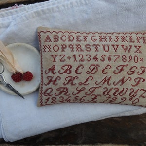 Pode incluir: Um pequeno travesseiro de cor creme com um alfabeto e números bordados em vermelho. O travesseiro está sobre um tecido de linho branco.