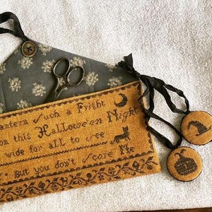 May include: A mustard yellow cross-stitch sampler with black text reading "Such a Fright on this Halloween Night." The design includes a cat, moon, and decorative border. Two round ornaments with a witch hat and pumpkin are also visible.