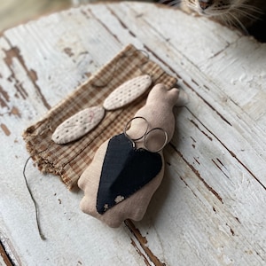 May include: A small, beige fabric bunny with a black heart sewn on its chest. The bunny is holding a pair of silver scissors. The bunny is sitting on a white wooden table.