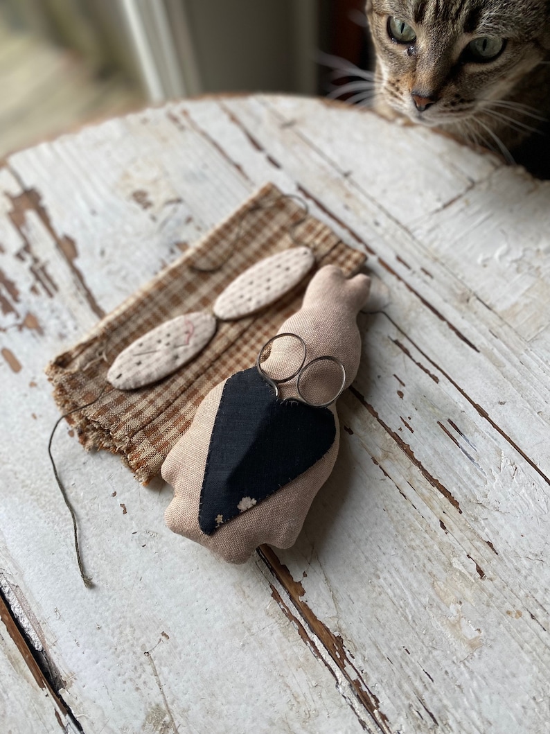 May include: A small, beige fabric bunny with a black heart sewn on its chest. The bunny is holding a pair of silver scissors. The bunny is sitting on a white wooden table.