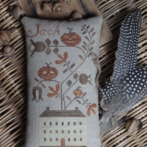 May include: A beige pillow with a cross-stitch design of a house with a pumpkin tree and the word "Jack" above it. The pillow is on a woven wicker surface with acorns and a feather.