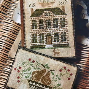 May include: Two cross-stitch embroidery pieces on a woven wicker background. The first piece features a brown house with a green roof and a flower pot with red flowers. The text "1820" is above the house. The second piece features a brown bowl with two pears and green leaves. Red berries are scattered around the bowl.