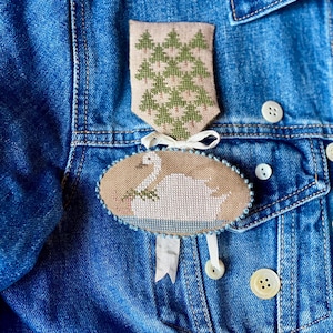 May include: A denim jacket adorned with two embroidered patches. The top patch features green trees on a beige background. The bottom patch is oval, depicting a white swan on a lake. Both patches are attached with white ribbons.