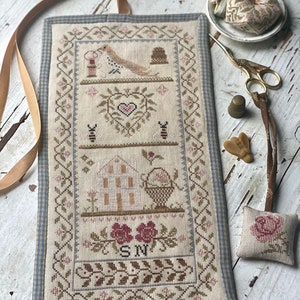 Op de afbeelding: Een naaldwerkproject met een blauwe en witte gingham rand. Het ontwerp bevat een vogel, een hart, een bijenkorf en een mand met bloemen. De initialen "SN" zijn geborduurd in de rechter benedenhoek.