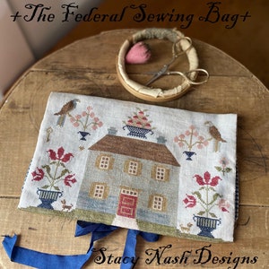 May include: A hand-stitched linen sewing bag with a blue ribbon closure. The bag features a cross-stitch design of a house with red tulips, birds, and a basket of fruit. The text "The Federal Sewing Bag" and "Stacy Nash Designs" are visible on the bag.