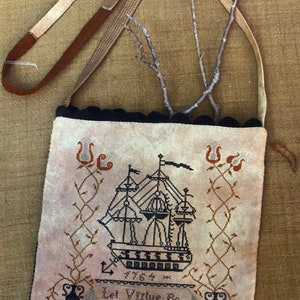 May include: A beige fabric bag with a black scalloped trim and a brown leather strap. The bag features an embroidered design of a ship with the text "Let Virtue Be A Guide to Thee" and the year "1764".