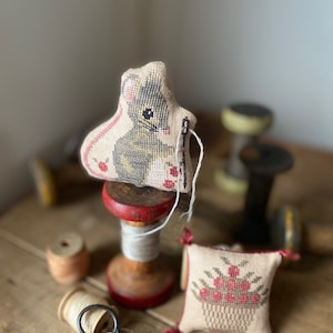 May include: A small, beige needle minder with a cross-stitch design of a mouse holding a needle. The needle minder is sitting on a wooden spool with red trim. There is a small, beige pincushion with a cross-stitch design of a basket of flowers. The pincushion is sitting on a wooden table with a pair of scissors.