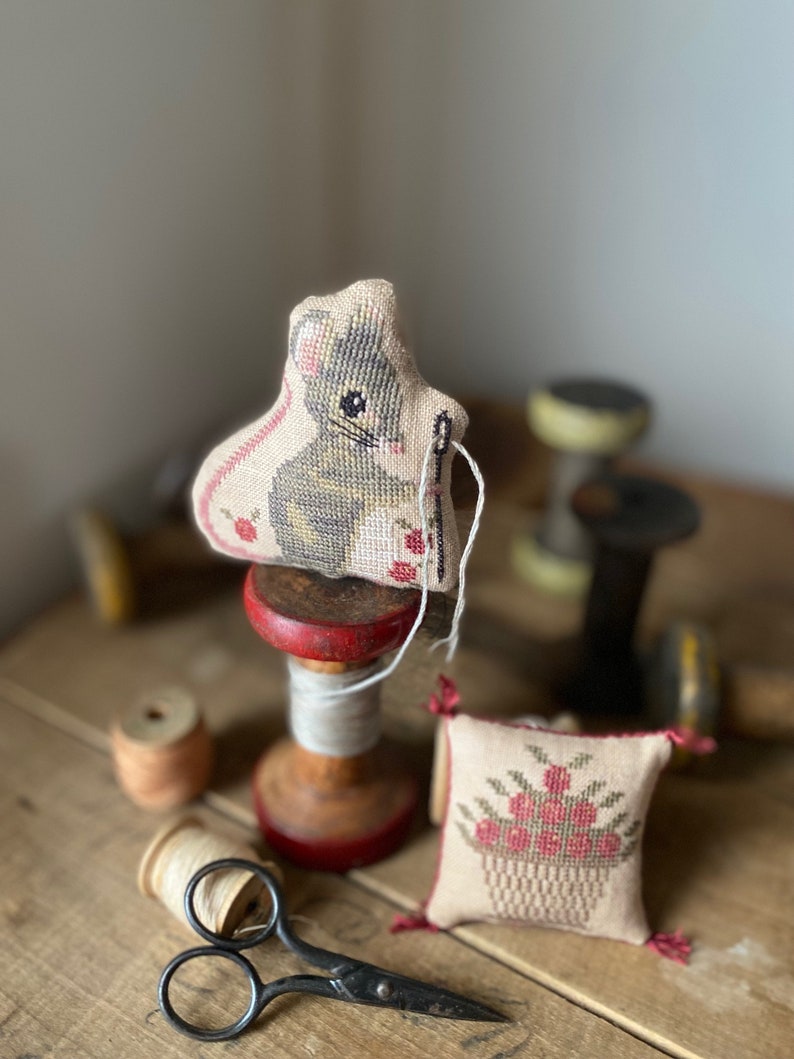 May include: A small, beige needle minder with a cross-stitch design of a mouse holding a needle. The needle minder is sitting on a wooden spool with red trim. There is a small, beige pincushion with a cross-stitch design of a basket of flowers. The pincushion is sitting on a wooden table with a pair of scissors.