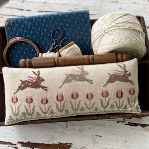 Op de afbeelding: Een rechthoekig beige kussen met een kruissteekontwerp van drie bruine konijnen die over rode tulpen springen. Het kussen ligt in een houten dienblad met garen, stof en naai-attributen.