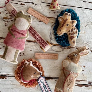 K&ouml;nnte beinhalten: Eine Sammlung handgefertigter Stofftier-Ornamente, darunter ein Frosch, eine Giraffe, ein Kaninchen und ein Igel. Jedes Ornament ist aufwendig mit verschiedenen Farben und Mustern bestickt. Holzlineale mit Text sind ebenfalls vorhanden, zusammen mit einer Partyhut.