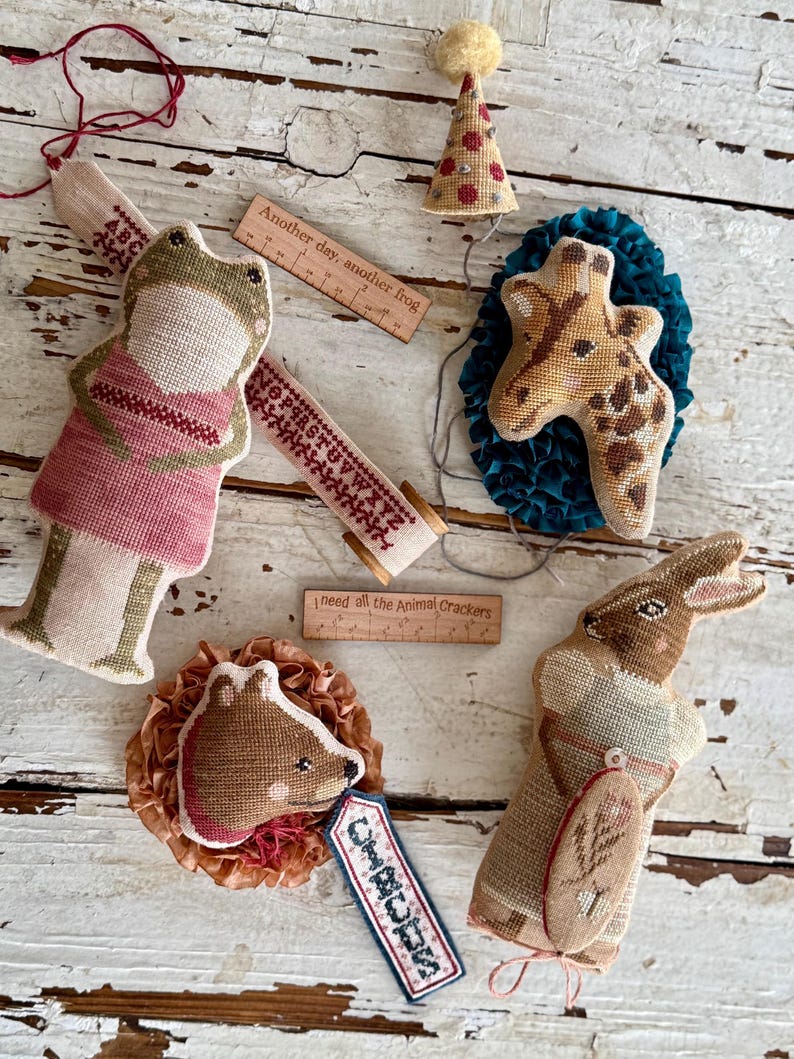 K&ouml;nnte beinhalten: Eine Sammlung handgefertigter Stofftier-Ornamente, darunter ein Frosch, eine Giraffe, ein Kaninchen und ein Igel. Jedes Ornament ist aufwendig mit verschiedenen Farben und Mustern bestickt. Holzlineale mit Text sind ebenfalls vorhanden, zusammen mit einer Partyhut.