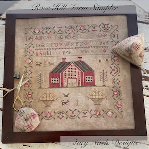 May include: A framed cross-stitch sampler, titled "Rose Hill Farm Sampler", featuring a red barn, floral border, and alphabet. Includes gold scissors, a thimble, and two pin cushions. The text "Stacy Nash Designs" is at the bottom.