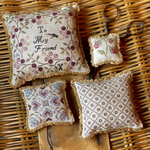 May include: Four small, square, embroidered throw pillows with fringe trim. The pillows are on a woven wicker surface. One pillow features a floral design with the text "To My Friend".