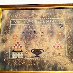 May include: A framed sampler with a faded, gray background. The sampler features a floral design with a vase, two baskets of fruit, and a small house. The alphabet is stitched in black thread at the top of the sampler. The sampler is signed "Mary Bovee".