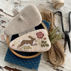 May include: A brown bunny embroidered on a cream-colored fabric with a red flower and green leaves. The fabric is in a wooden embroidery hoop and is sitting on top of a blue book. There is a spool of twine and a pair of scissors in the background.