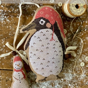 May include: Cross-stitched penguin and snowman ornaments. The penguin has a red cap and a white belly with black accents. The snowman wears a red hat and scarf. The text "Animal Cracker Series + Chilli + Stacy Nash Designs" is visible.