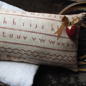 May include: A rectangular, beige embroidered pillow with red stitching featuring the alphabet, decorative lines, and the name "Harriet Brown" and the year "1837". A red strawberry pin with a gold ribbon is attached to the pillow.