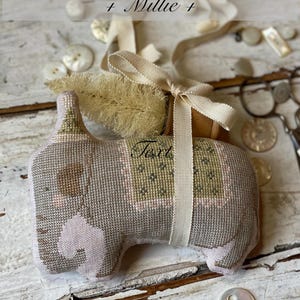 May include: A handcrafted elephant-shaped ornament from the "Animal Cracker Series + Millie +" collection. The ornament is mainly grey with pink details and a green and tan patterned saddle. It is tied with a cream ribbon and a small decorative brush.