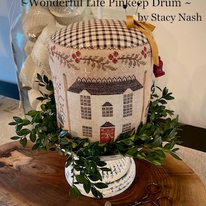 Puede incluir: Un cojín para alfileres con forma de casa con una puerta roja y ventanas blancas. El cojín está decorado con una tela a cuadros marrón y blanca y una corona de hojas verdes. El cojín está sentado sobre un pedestal blanco y una superficie de madera. El texto "Wonderful Life Pinkeep Drum ~ by Stacy Nash" es visible en la imagen.