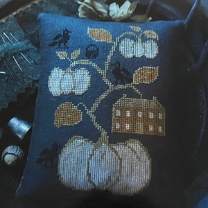 以下が含まれることがあります： 青い針仕事用のクッションで、家、カボチャ、鳥の刺繍が施されています。クッションの周りは、針、指ぬき、糸巻きなど、裁縫道具で囲まれています。