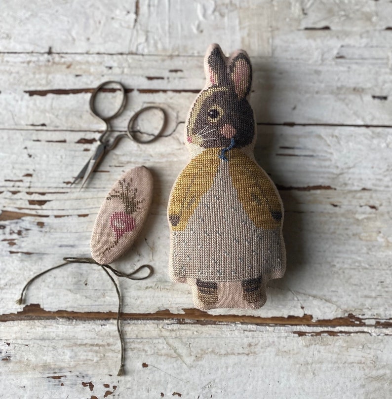 May include: A brown and white embroidered bunny doll wearing a yellow jacket. The doll is sitting on a white wooden surface with a small embroidered piece of fabric and a pair of scissors.