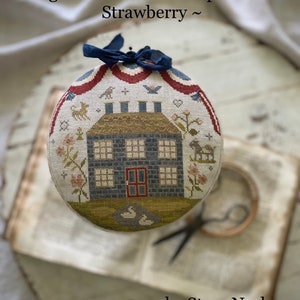 Puede incluir: Un proyecto de bordado de punto de cruz terminado que representa una casa azul con molduras rojas y una valla blanca. La casa está rodeada de flores y pájaros. El título del proyecto es "Big Blue House Pinkeep Drum & Strawberry" de Stacy Nash.