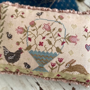 Op de afbeelding: Een rechthoekig, kruissteek kussen met een lichtroze achtergrond en een gerimpelde rand. Het ontwerp toont een bloemenmand, een kip, een konijn en kleine vogels. Het kleurenpalet omvat roze, blauw, groen en bruin.