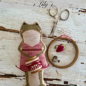K&ouml;nnte beinhalten: Ein handgemachtes froschf&ouml;rmiges Nadelkissen mit einem rosa Kleid und einer roten Garnrolle. Das Bild zeigt auch einen kleinen Stickrahmen mit einer Erdbeere, einem Fingerhut und einer Schere. Der Text lautet "Animal Cracker Series + Lily + Stacy Nash Designs."