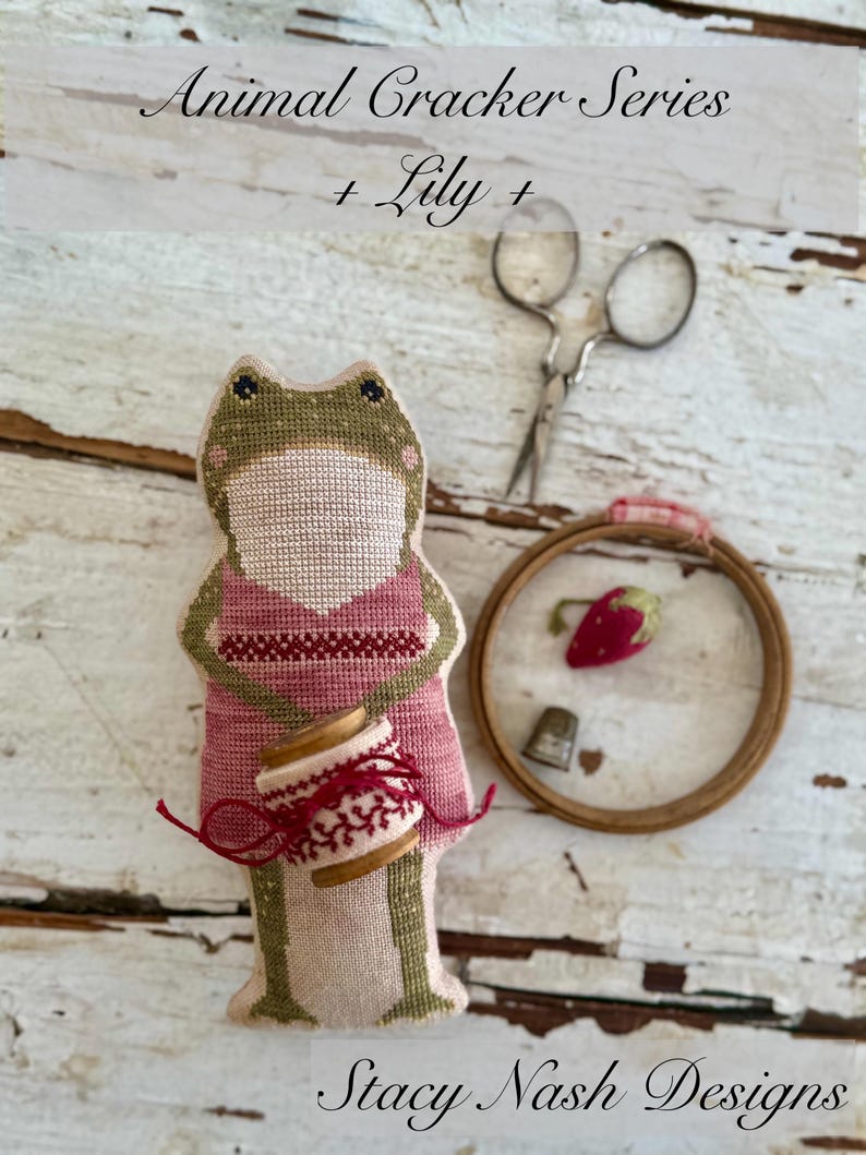 K&ouml;nnte beinhalten: Ein handgemachtes froschf&ouml;rmiges Nadelkissen mit einem rosa Kleid und einer roten Garnrolle. Das Bild zeigt auch einen kleinen Stickrahmen mit einer Erdbeere, einem Fingerhut und einer Schere. Der Text lautet "Animal Cracker Series + Lily + Stacy Nash Designs."