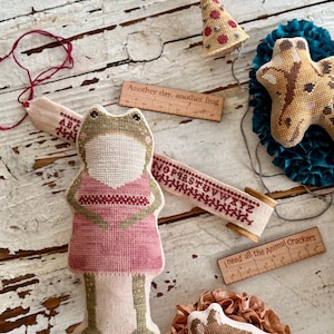 K&ouml;nnte beinhalten: Eine Sammlung handgefertigter Stoffornamente und Accessoires. Enth&auml;lt ein froschf&ouml;rmiges Ornament in einem rosa Kleid, eine Giraffe, eine Partyhut und einen Igel. Holzlineale mit Text: "Another day, another frog" und "I need all the Animal Crackers".