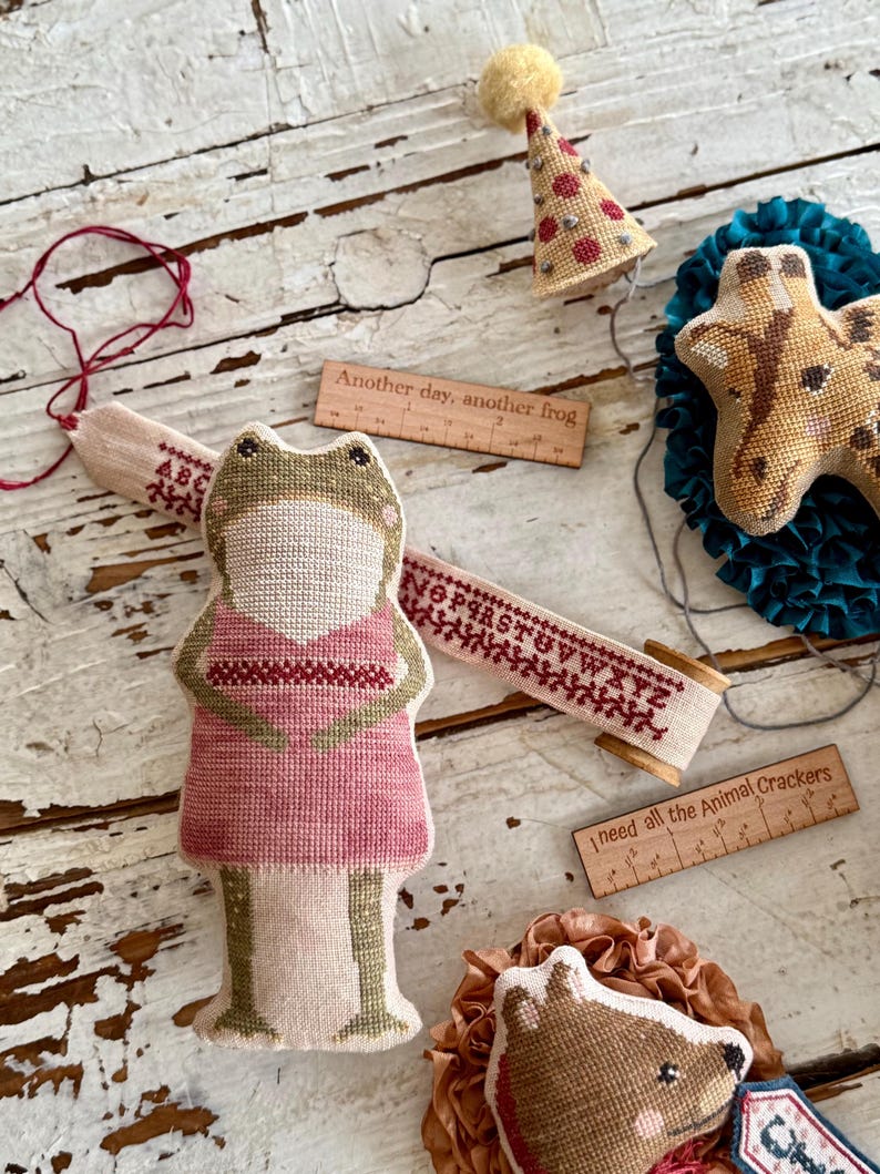 K&ouml;nnte beinhalten: Eine Sammlung handgefertigter Stoffornamente und Accessoires. Enth&auml;lt ein froschf&ouml;rmiges Ornament in einem rosa Kleid, eine Giraffe, eine Partyhut und einen Igel. Holzlineale mit Text: "Another day, another frog" und "I need all the Animal Crackers".