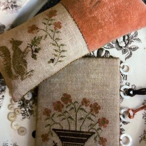 Könnte beinhalten: Zwei gestickte Nadelfilzkissen mit Blumendesigns. Ein Kissen ist hellbraun mit einem grünen Blumendesign und einem Eichhörnchen. Das andere Kissen ist hellbraun mit einem braunen Blumendesign in einem Topf. Die Kissen liegen auf einem weißen Hintergrund mit einem schwarz-weißen Blumenmuster.