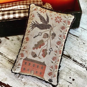 Könnte beinhalten: Rechteckiges Zierkissen mit detailliertem Kreuzstichdesign. Das Kissen zeigt einen braunen Vogel, ein rotes Haus, Schlüssel und florale Elemente. Das Kissen hat einen schwarzen Rüschenrand und liegt auf einer rustikalen Holzoberfläche.