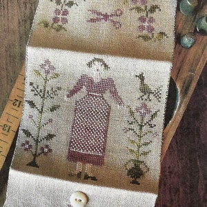 May include: A cross-stitch sampler with a woman in a purple dress standing between two plants. The sampler is folded in thirds and has a white button on the bottom section.