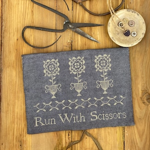 May include: A gray fabric sampler with white embroidery of three stylized flower pots and the text "Run With Scissors". The sampler is on a wooden surface with two pairs of scissors and a small ceramic dish with buttons.