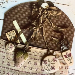 May include: A brown and beige fabric needlepoint project bag with a brown and white plaid flap. The bag has a brown ribbon tied around it and a button. The bag is decorated with a variety of needlepoint designs, including a strawberry, a flower, and a pair of scissors. The bag also has a needlepoint alphabet and numbers.