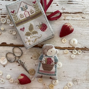 Può includere: Una collezione di oggetti fatti a mano a punto croce, tra cui un astuccio per aghi a forma di casa, una figura di riccio con un cestino di fragole e due cuscini a forma di fragola. Piccoli bottoni bianchi e un paio di forbici sono sparsi intorno agli oggetti. Il testo "Animal Cracker Series + Pip+" e "Design by Stacy Nash" sono visibili.