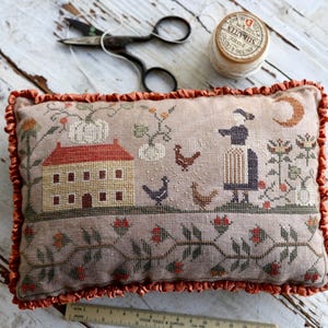 May include: A beige cushion with a brown, orange and white embroidered design of a house, pumpkins and chickens. The cushion has a ruffled orange trim.