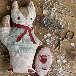 May include: A handcrafted fox Christmas decoration wearing a red scarf and a green sweater. An oval ornament with a hot chocolate design is also present. The scene includes snowflake decorations, buttons, and small scissors on a wooden surface.