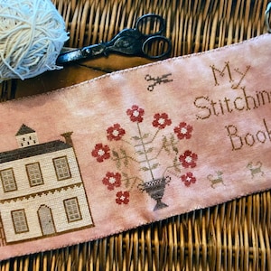 Puede incluir: Una aguja de punto de tela rosa con una casa, flores en un jarrón y el texto "My Stitching Book".