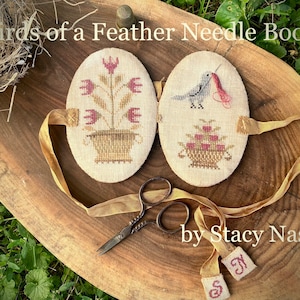 May include: A needle book with a floral design on one side and a bird with a needle in its beak on the other side. The book is made of white fabric and has a yellow ribbon tied around it. The text "Birds of a Feather Needle Book" is written above the book. The text "by Stacy Nash" is written below the book.
