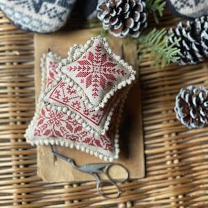 Puede incluir: Dos pequeños cojines cuadrados bordados en rojo y blanco con un diseño de copo de nieve y pompones blancos alrededor de los bordes. Los cojines están apilados uno encima del otro sobre una superficie marrón.