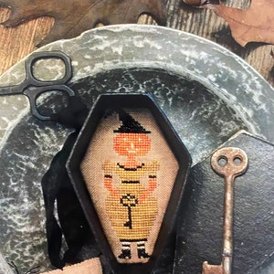Pode incluir: Uma moldura preta em forma de caixão com um desenho de ponto cruz de uma bruxa com cara de abóbora segurando uma chave. A bruxa está usando um chapéu preto e um vestido amarelo com listras pretas. A moldura está sobre uma superfície de metal cinza com uma chave e um pequeno ponto cruz de uma abóbora.
