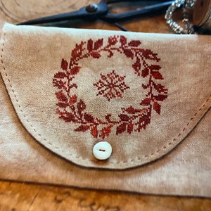May include: A small, beige fabric pouch with a red embroidered wreath and snowflake design. The pouch has a white button closure.