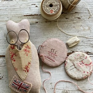 Puede incluir: Un coj&iacute;n de alfileres de tela beige con un dise&ntilde;o floral y un peque&ntilde;o parche de la Union Jack. El coj&iacute;n de alfileres est&aacute; lleno de agujas e hilo. Tambi&eacute;n hay dos carretes de hilo, uno con una etiqueta que dice "Knitting & Crochet Pure Sixteen 125 Yds Fall River K.T.Co."